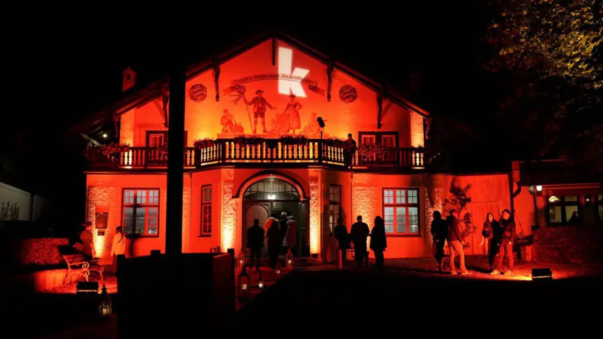 Beleuchtetes Gebäude bei Nacht in rotem Licht mit Balkon und Eingang, davor mehrere Personen; an der Fassade ein großes beleuchtetes K.