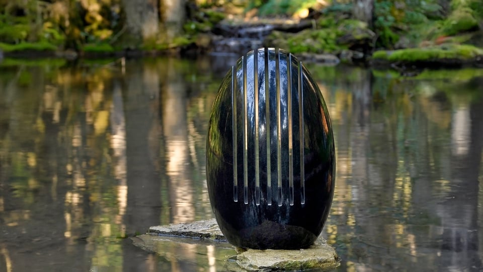Skulptur eines schwarzen, ovalen Klangsteins mit vertikalen Metallstreifen auf einem Stein im Wasser, umgeben von einem ruhigen Waldteich.