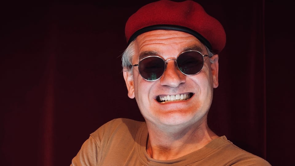 Person mit roter Baskenmütze und runder Sonnenbrille vor dunkelrotem Hintergrund. Die Person grinst weit, zeigt dabei ihre Zähne und trägt ein hellbraunes T Shirt.