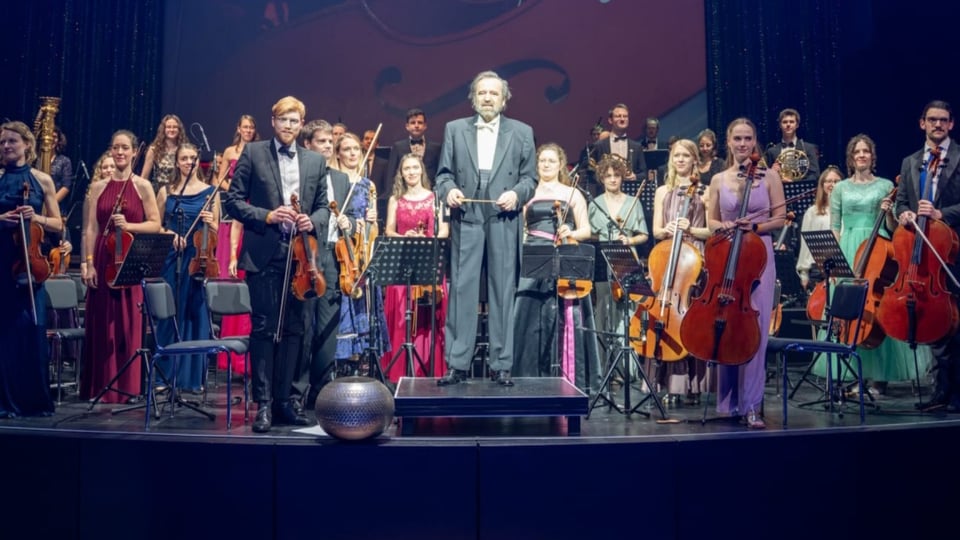 Orchester auf einer Konzertbühne mit Dirigent auf dem Podium. Rundherum stehen mehrere junge Musikerinnen und Musiker mit Violinen, Violoncelli und weiteren Orchesterinstrumenten in festlicher Konzertkleidung vor ihren Notenständern.