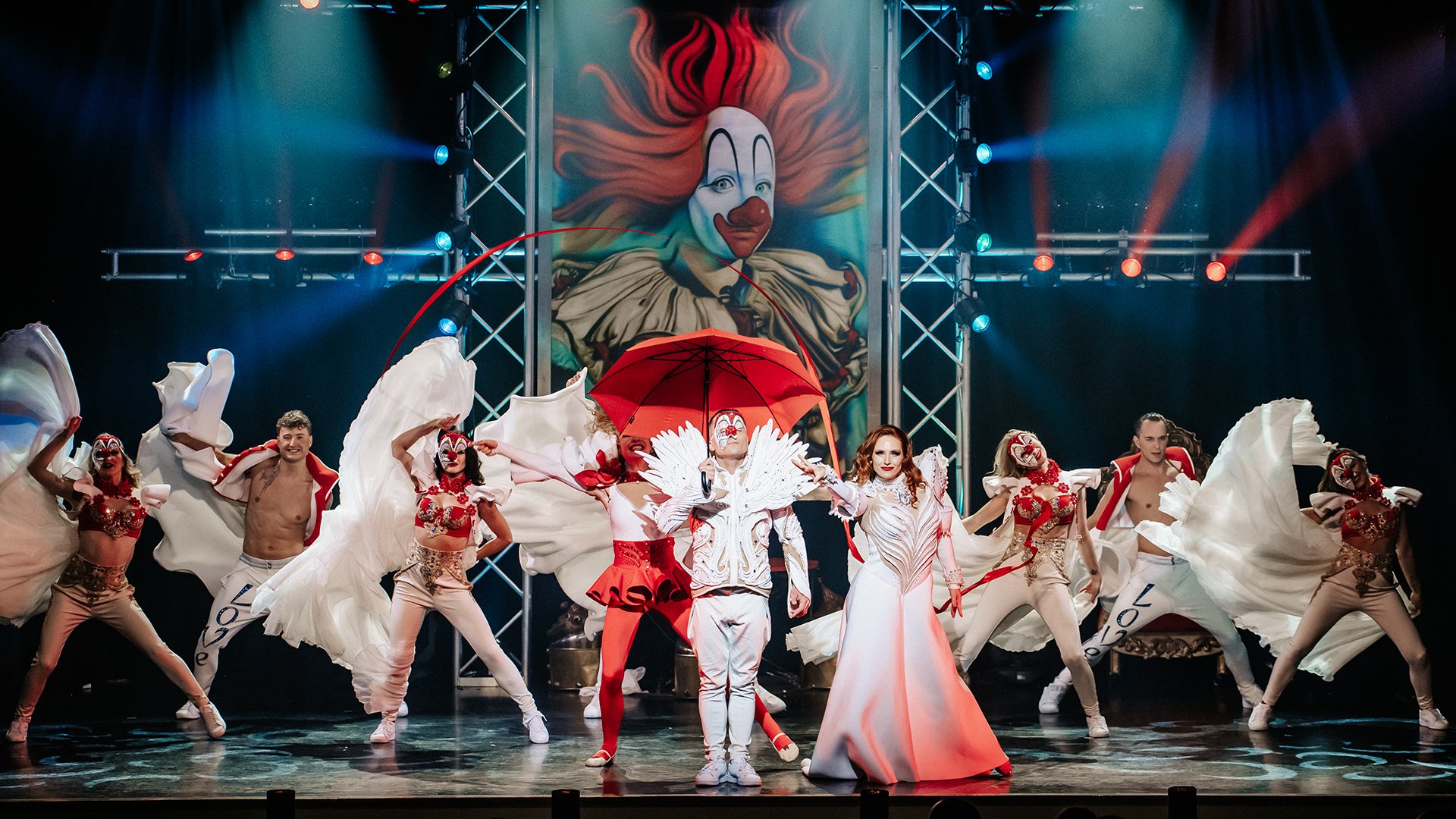 Bühnenfoto aus einer GOP Varieté-Show mit mehreren Artistinnen und Artisten in weißen und roten Kostümen auf einer beleuchteten Bühne, im Zentrum eine Person mit rotem Schirm, im Hintergrund ein großes Clownsmotiv und farbige Lichteffekte