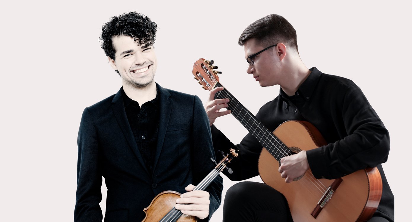 Die beiden Künstler sind vor einem weißen Hintergrund mit Gitarre und Geige zu sehen und lachen fröhlich