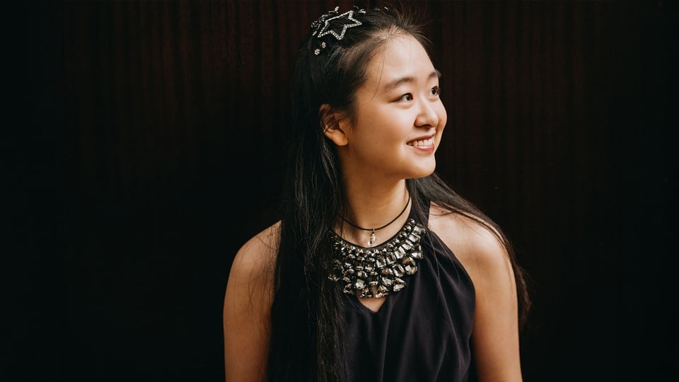 Tianyao Lyu mit langen dunklen Haaren vor dunklem Hintergrund, trägt ein schwarzes Oberteil mit auffälligem Schmuck am Ausschnitt und blickt seitlich.