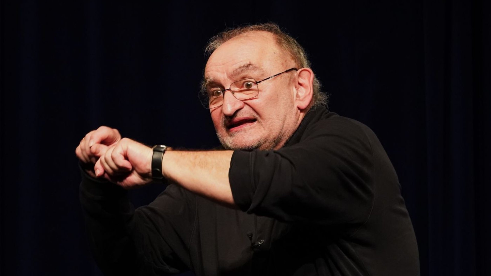 Sigi Zimmerschied mit Brille und schwarzem Hemd steht auf einer Bühne vor dunklem Hintergrund und zeigt mit gestikulierenden Armen eine ausdrucksstarke Geste während eines Kabarettauftritts.