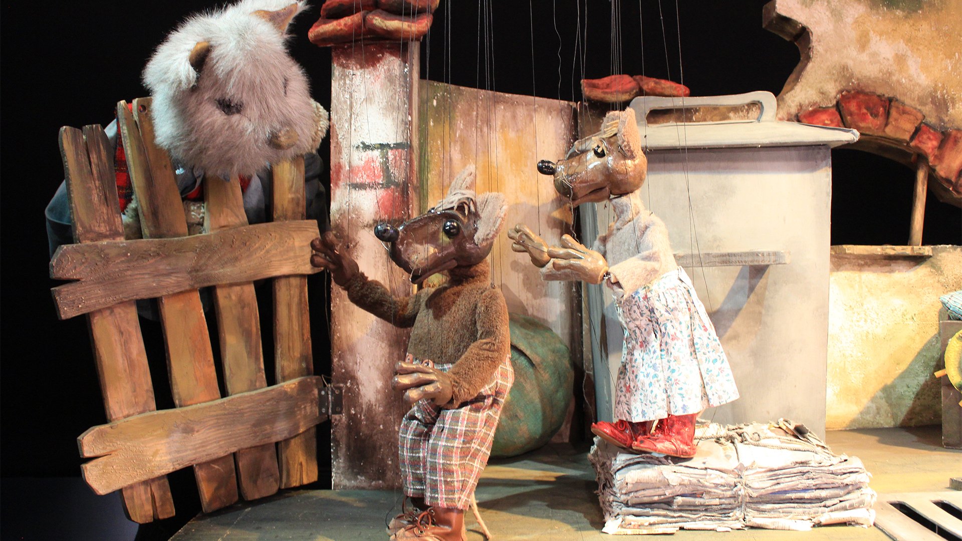 Puppenszene mit zwei aufrecht stehenden Rattenfiguren vor einer Mauer und einem großen Metallbehälter; links schaut ein großes, weißes Tier durch einen hölzernen Zaun, während auf dem Boden Papierstapel und weitere Requisiten der Bühnenkulisse liegen.