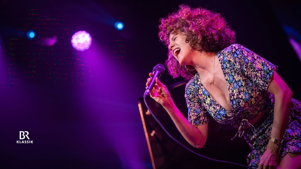 Foto vom Event von Cyrille Aimée mit der Künstlerin mit lockigem Haar, die mit einem Mikrofon singt