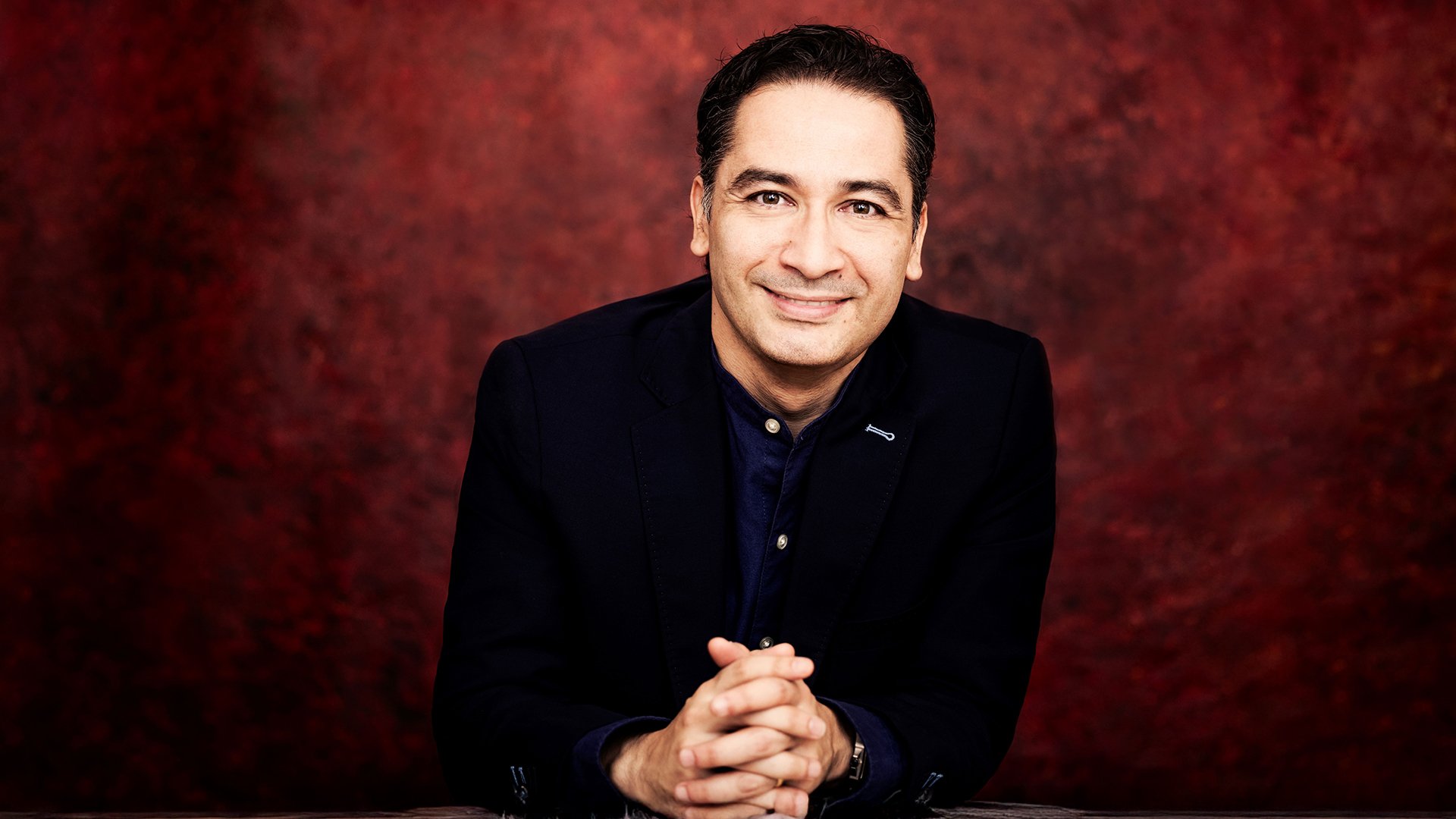 Andres Orozco in dunkler Kleidung, mit gefalteten Händen vor rotem Hintergrund.