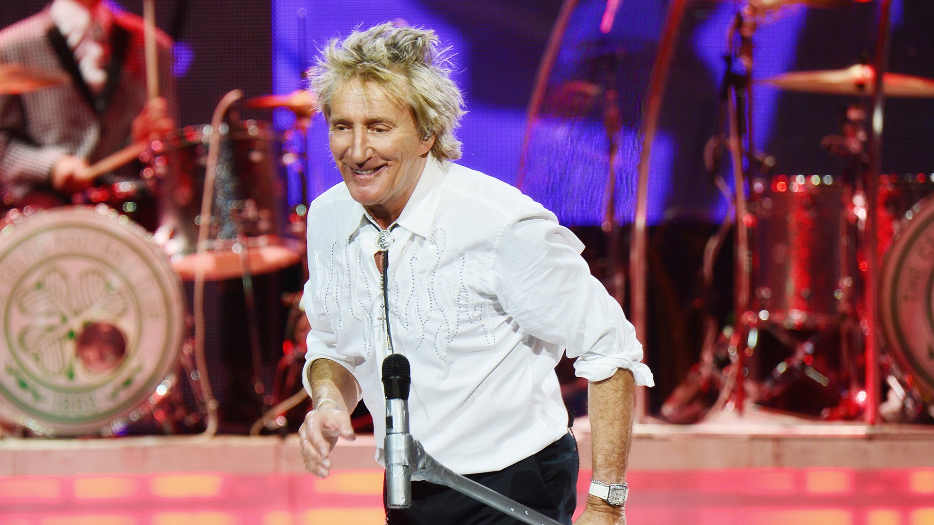 Bühnenfoto von Rod Stewart in weißem Hemd mit Mikrofon auf einer Konzertbühne, vor Schlagzeug und rot violett beleuchteter Showkulisse.
