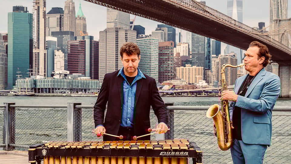 Eventfoto von Mulo Francel & Tim Collins mit beiden Künstlern – einer mit Saxofon, der andere mit Xylophon – vor einer Skyline mit Wolkenkratzern