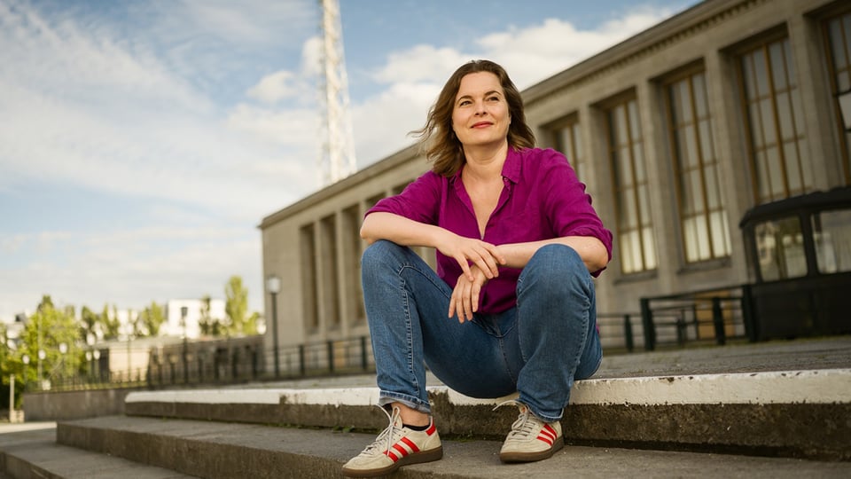 Katja Berlin sitzt draußen auf einer Treppe vor einem Gebäude und lächelt.