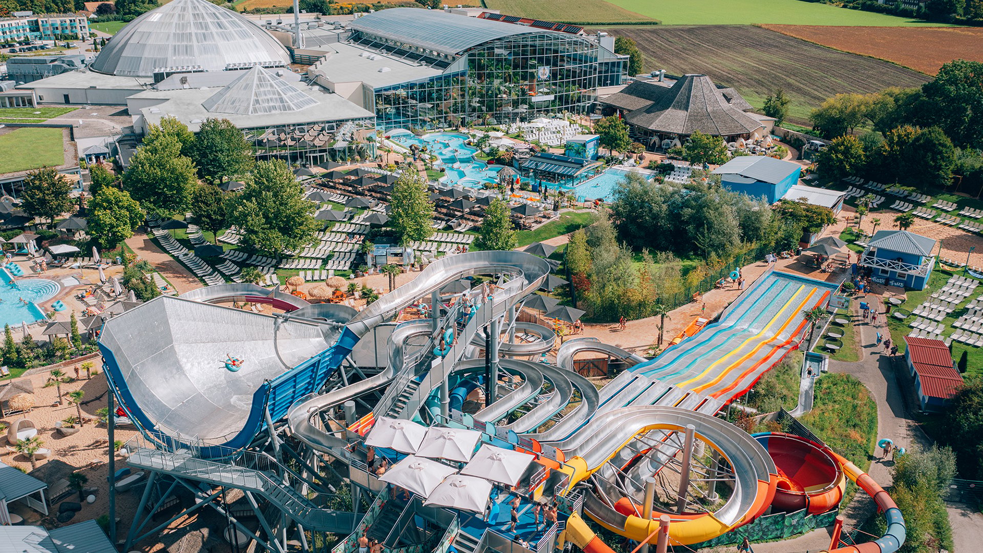 Luftaufnahme der Therme Erding mit großen Glasdächern, Außenpools, vielen Liegen sowie mehreren bunten Wasserrutschen und Rutschenanlagen im Vordergrund