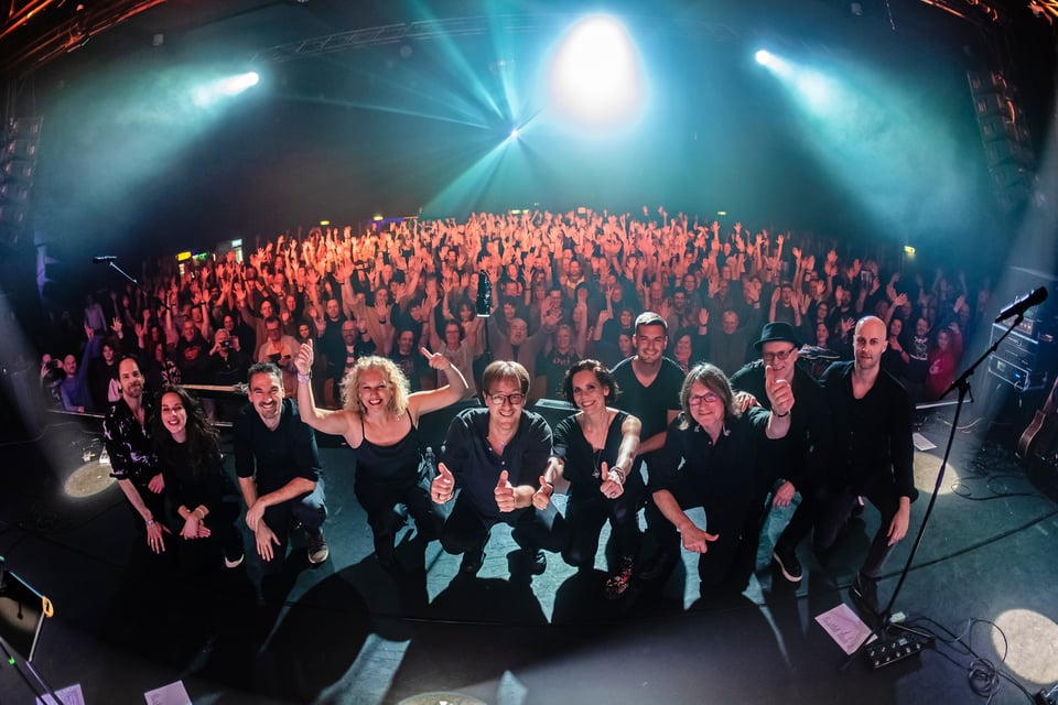 Bühnenfoto von Floyd Division mit neun Bandmitgliedern am Bühnenrand vor ausverkauftem Publikum, die gemeinsam in die Kamera blicken, während zahlreiche Konzertbesuchende im Hintergrund die Hände heben, unter blauem Bühnenlicht und Scheinwerfern.