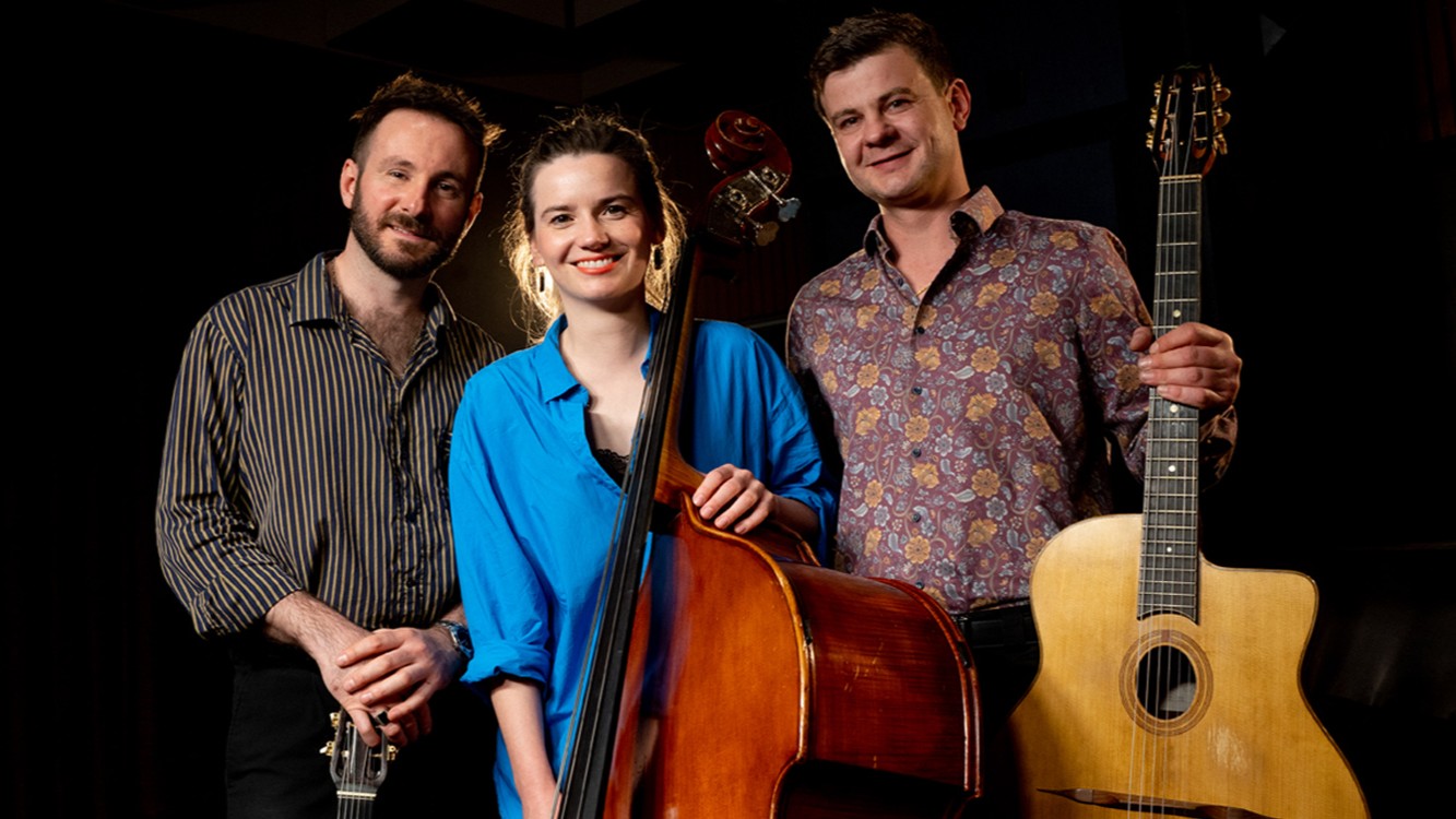 Die European Django Band mit drei Musikerinnen und Musikern vor dunklem Hintergrund, eine Person mit Kontrabass in blauem Hemd in der Mitte, flankiert von zwei Personen mit Gitarren.