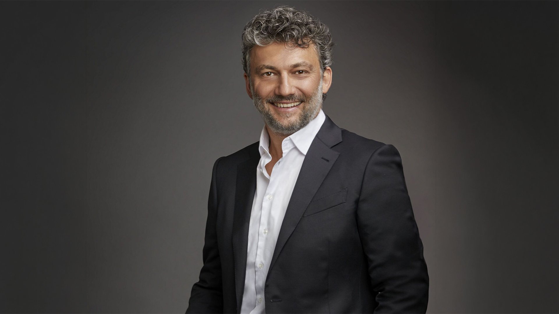 Porträtfoto von Jonas Kaufmann vor neutralem grauem Hintergrund in dunklem Anzug und weißem Hemd.