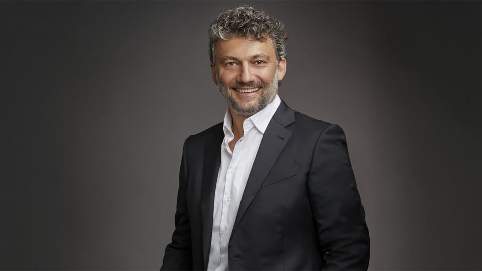 Porträtfoto von Jonas Kaufmann vor neutralem grauem Hintergrund in dunklem Anzug und weißem Hemd.