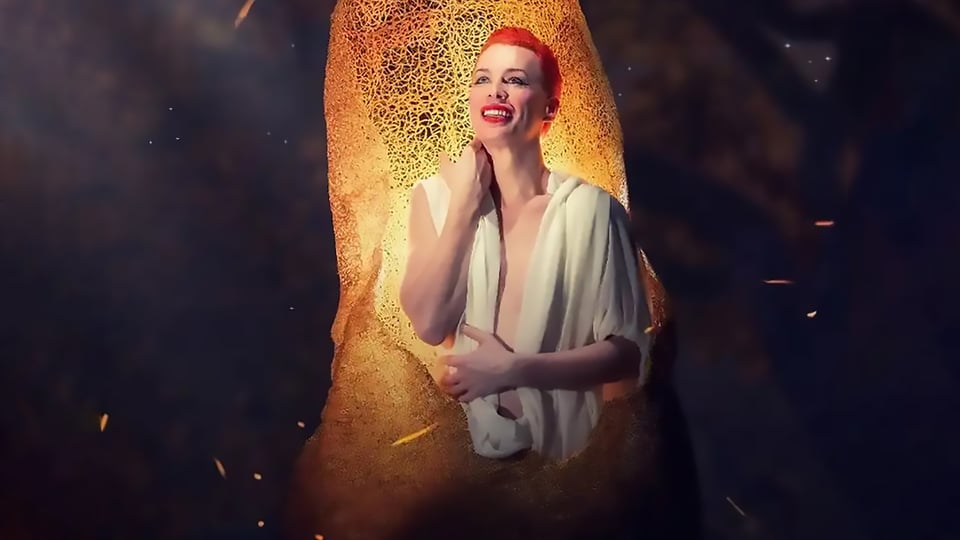 Die Künstlerin DIVINA DE CAMPOmit kurzen roten Haaren und weißer Bluse sitzt in einem goldfarbenen Gebilde
