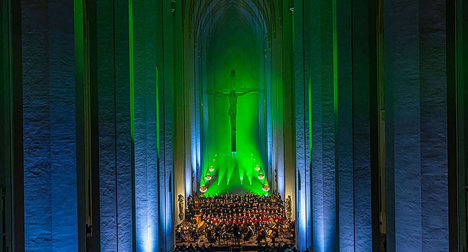 Foto eines Orchesters in einer Kirche mit einem riesigen Christus am Kreuz und grüner Beleuchtung.