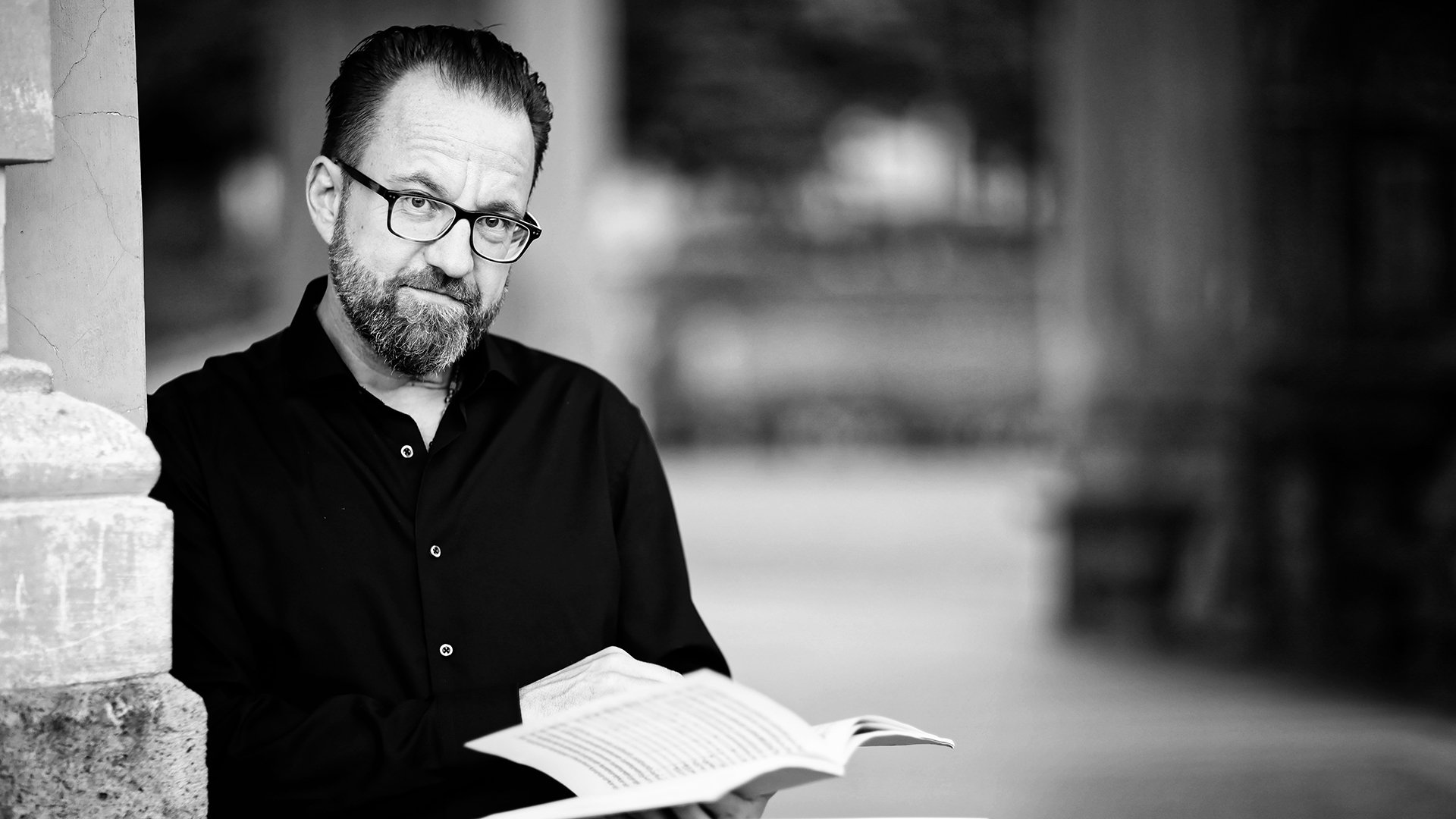 Schwarz-weiss Portrait vom Dirigenten Franz Schottky an einem Mauervorsprung lehnend mit einer Mappe in der Hand