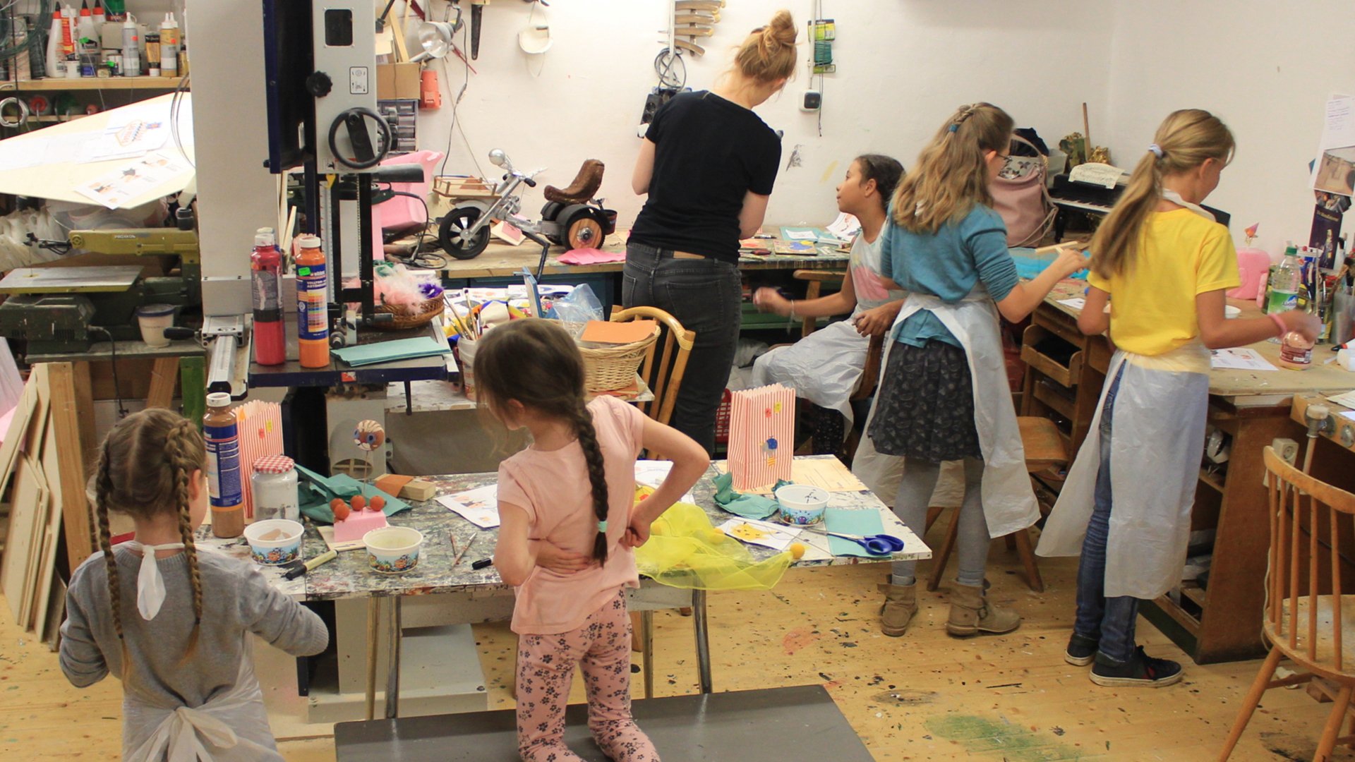Mehrere Kinder und eine erwachsene Person arbeiten in einer gut ausgestatteten Werkstatt an Bastel- und Kreativprojekten; auf den Tischen liegen Papier, Farben, Scheren und verschiedene Materialien, während die Kinder konzentriert schneiden, malen und gestalten.