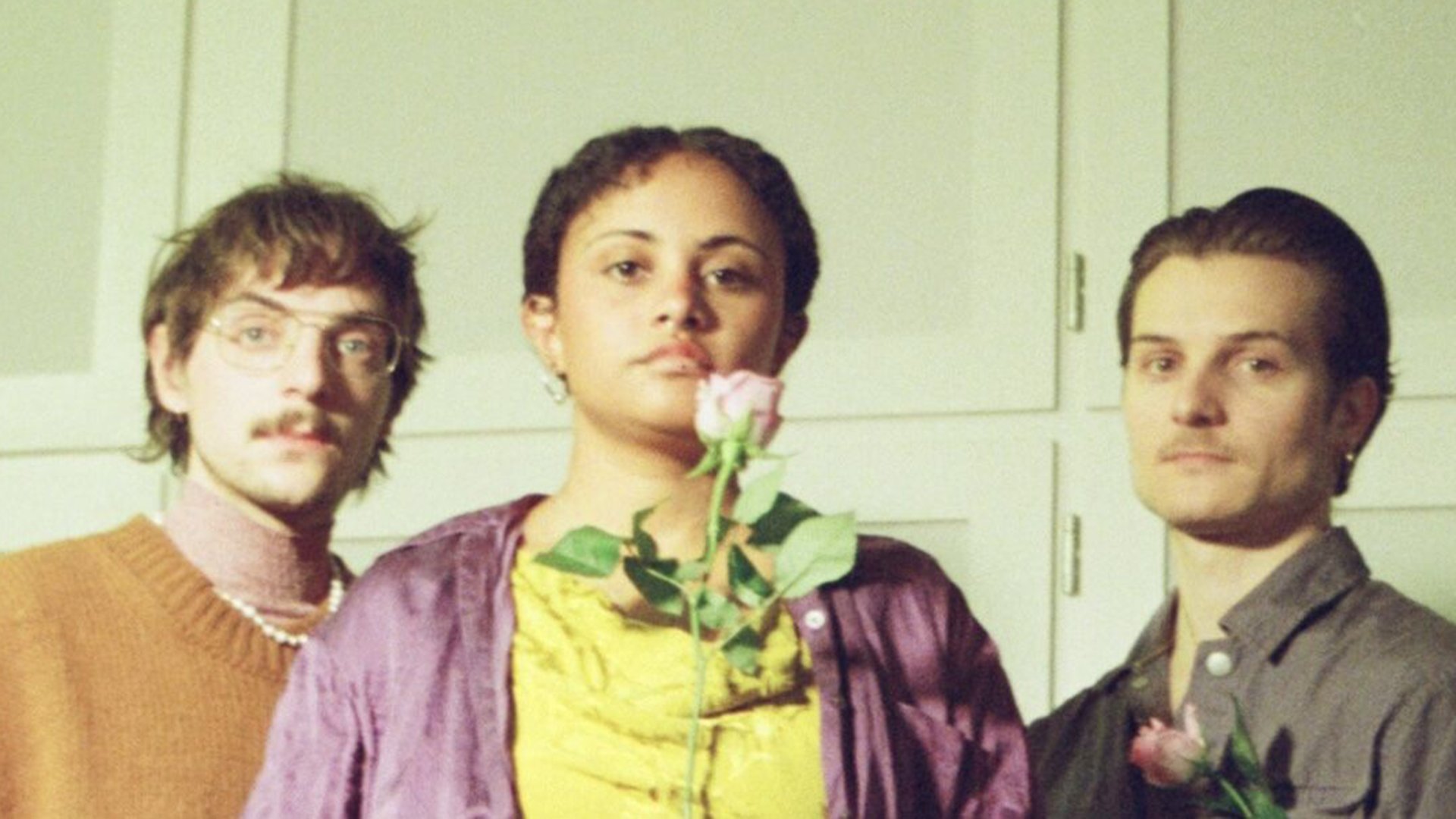 Drei Personen stehen nebeneinander vor hellem Hintergrund, blicken ruhig in die Kamera und halten jeweils eine Blume, warmes Licht und leicht nostalgischer, unscharfer Bildstil