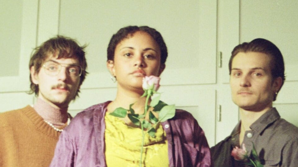 Drei Personen stehen nebeneinander vor hellem Hintergrund, blicken ruhig in die Kamera und halten jeweils eine Blume, warmes Licht und leicht nostalgischer, unscharfer Bildstil