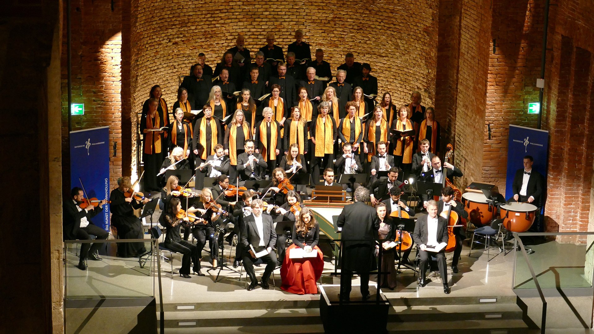 Bühnenfoto von Chor und Orchester bei einer Aufführung des Weihnachtsoratoriums in einem hohen Backsteinbau, mit Dirigent vor dem Ensemble, Solistinnen und Solisten im Vordergrund sowie Sängerinnen und Sängern mit goldfarbenen Schals auf gestaffelten Podesten im Hintergrund.
