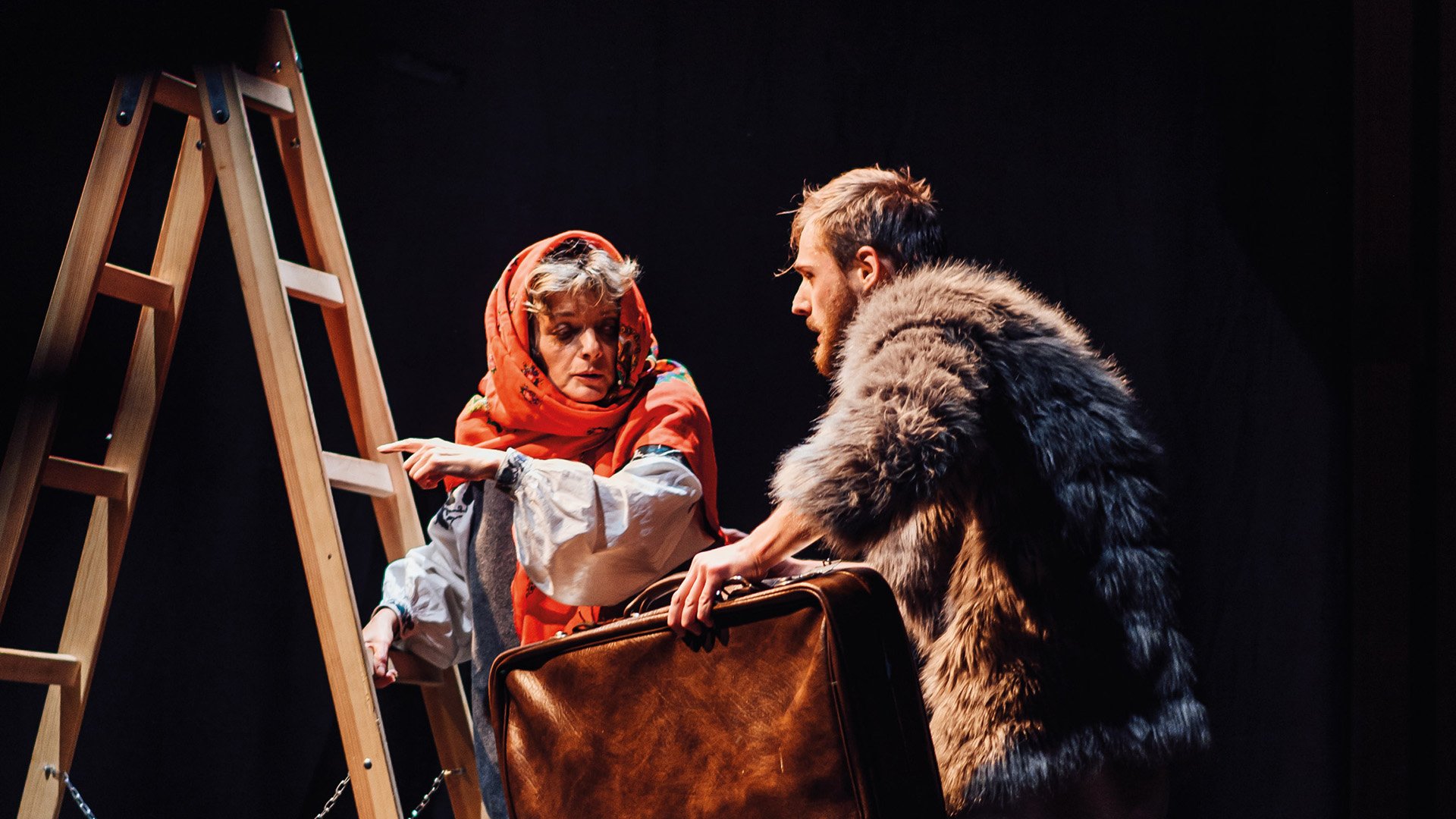 Zwei Personen in Kostümen stehen auf einer Theaterbühne neben einer Holzleiter. Eine Person trägt ein rotes Kopftuch und zeigt auf die Leiter, während die andere einen großen Koffer hält und einen pelzartigen Umhang trägt. Die Szene wird von warmem Bühnenlicht vor dunklem Hintergrund beleuchtet.