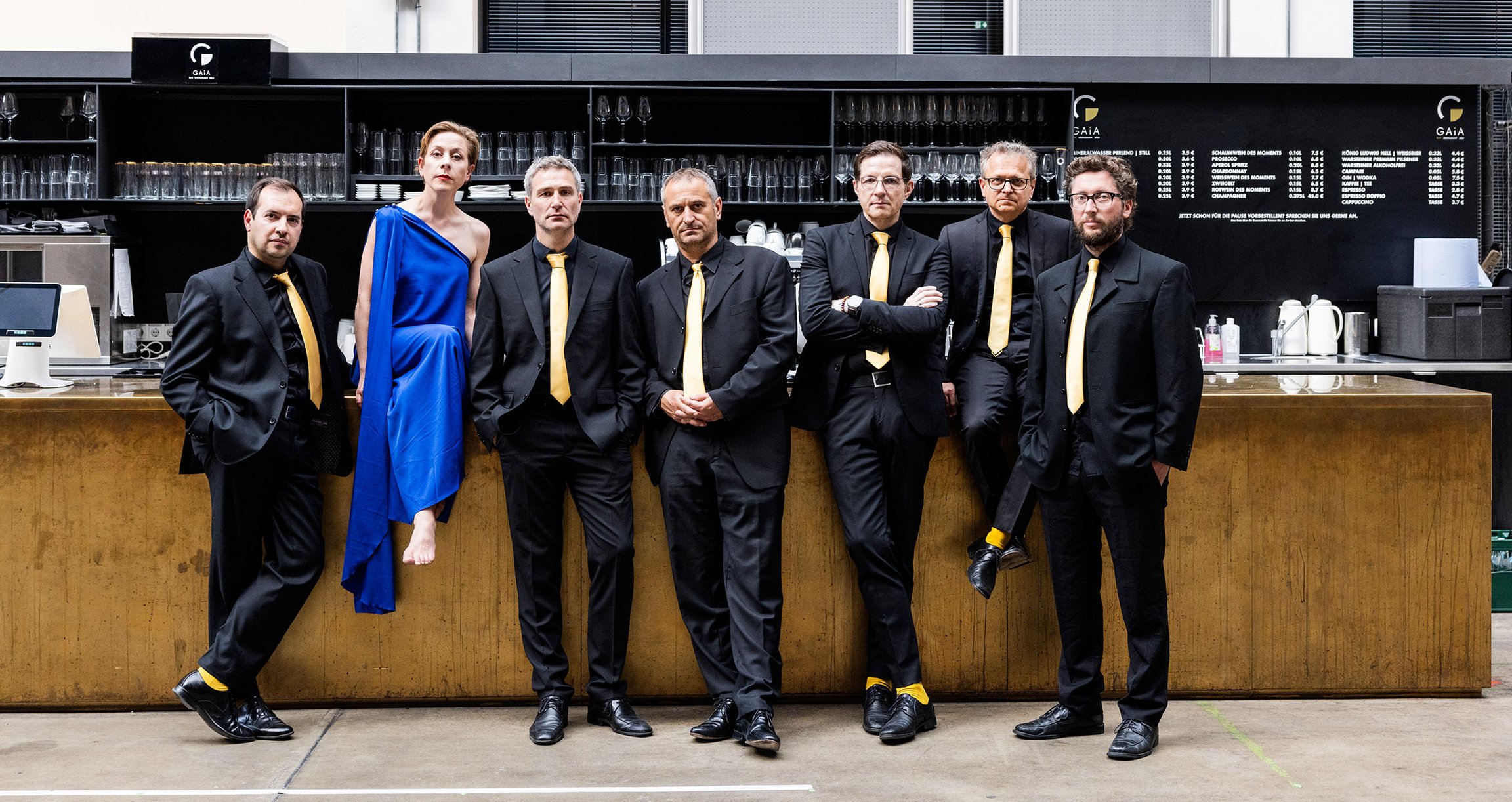 Sechs Herren in dunklen Anzügen und eine Dame in blauem Kleid vor Tresen stehend
