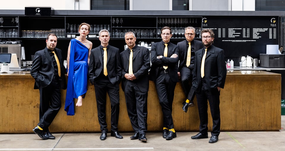Sechs Herren in dunklen Anzügen und eine Dame in blauem Kleid vor Tresen stehend