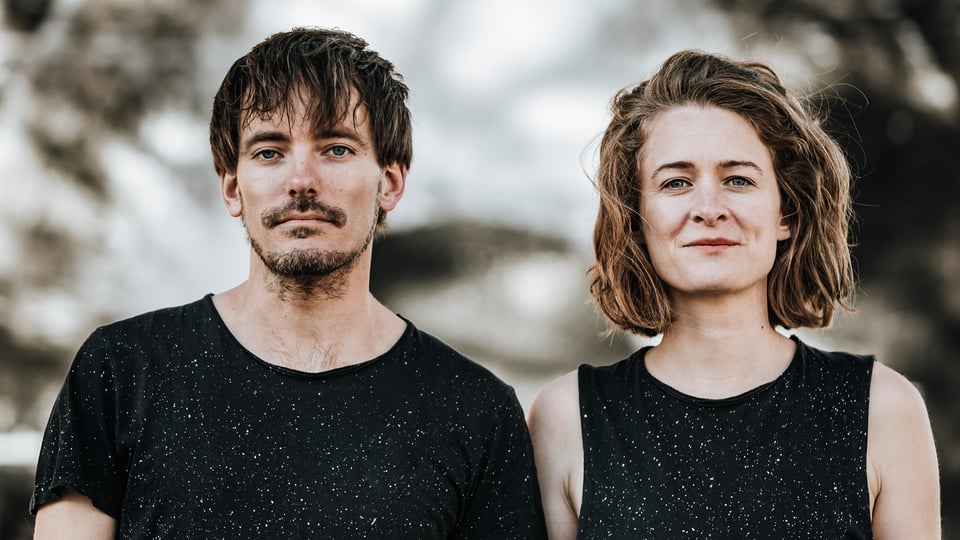 Zwei Personen stehen nebeneinander im Freien und tragen dunkle, kurzärmelige Oberteile mit feinen hellen Sprenkeln; im Hintergrund ist eine naturhafte, unscharfe Umgebung zu erkennen