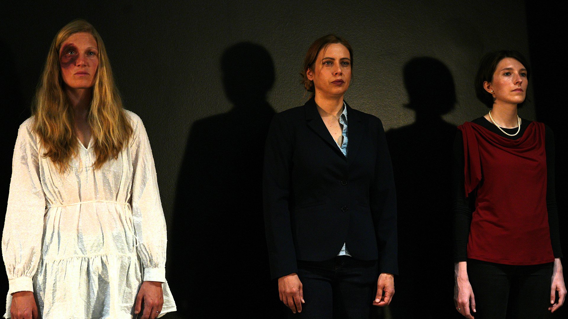 Bühnenfoto aus Libertà o quasi mit drei Darstellenden vor schwarzem Hintergrund, links eine Person in weißem Kleid mit dunklem Auge-Make-up, in der Mitte eine Person im dunklen Hosenanzug und rechts eine Person mit rotem Oberteil und Perlenkette, alle nebeneinander stehend im Scheinwerferlicht.