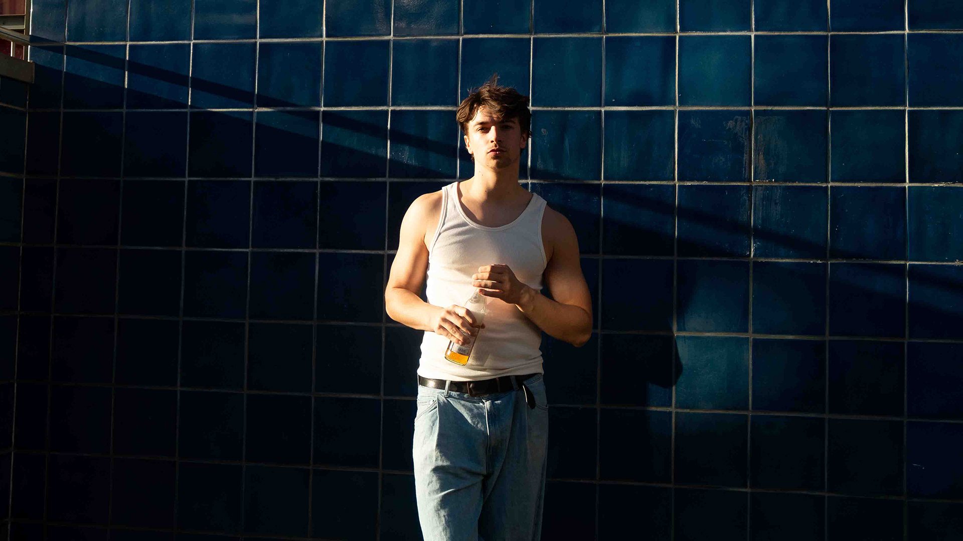 Mika Noe steht vor einer Wand aus blauen Fliesen und guckt mit einer Flasche Saft in die Kamera