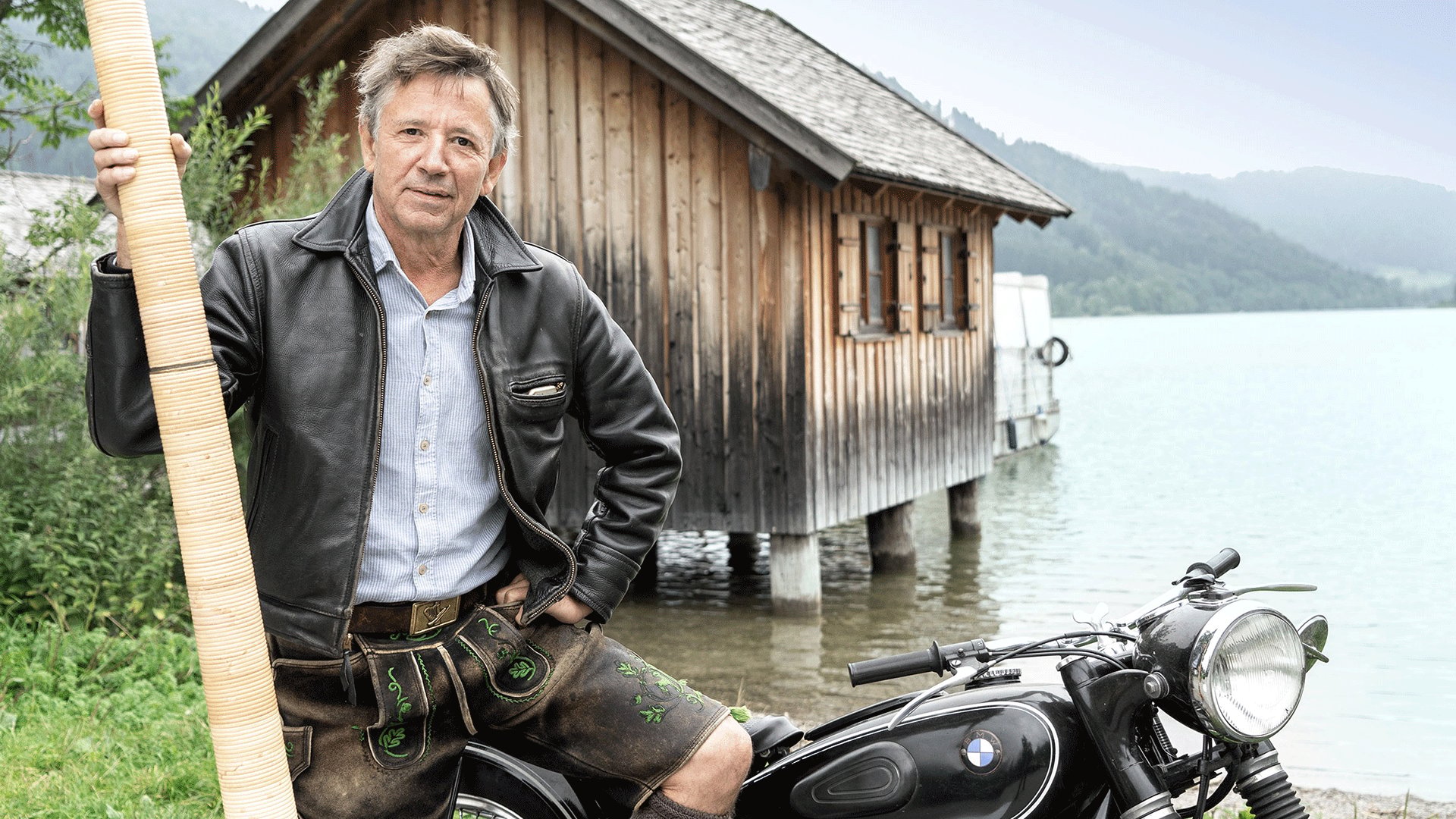 Person in Lederhose und Lederjacke neben einem historischen Motorrad am Ufer eines Sees und hält ein langes Alphorn. Im Hintergrund stehen ein Holzhaus auf Pfählen sowie bewaldete Berge am Seeufer.