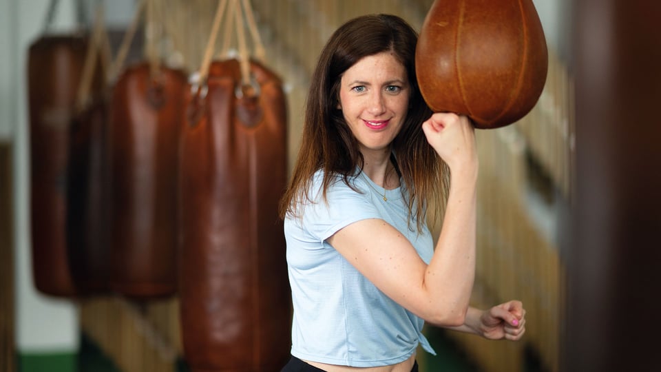 Claudia Pichler im hellblauen T Shirt, die mit erhobenem Arm einen Boxsack schlägt, in Trainingsraum mit mehreren braunen Boxsäcken im Hintergrund