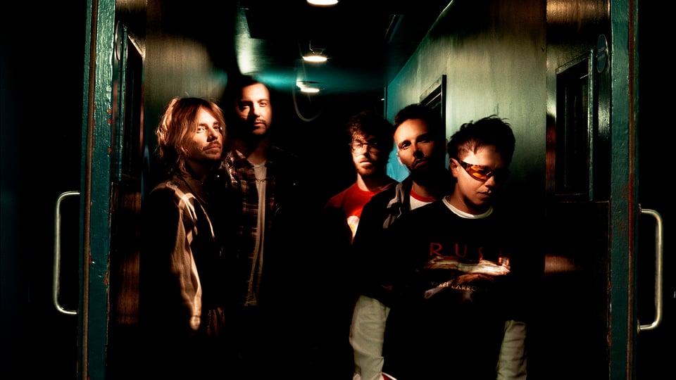 Bandfoto von fünf Musikern in einem dunklen, atmosphärisch beleuchteten Gang mit dramatischem Licht und Indie-Rock-Ästhetik.