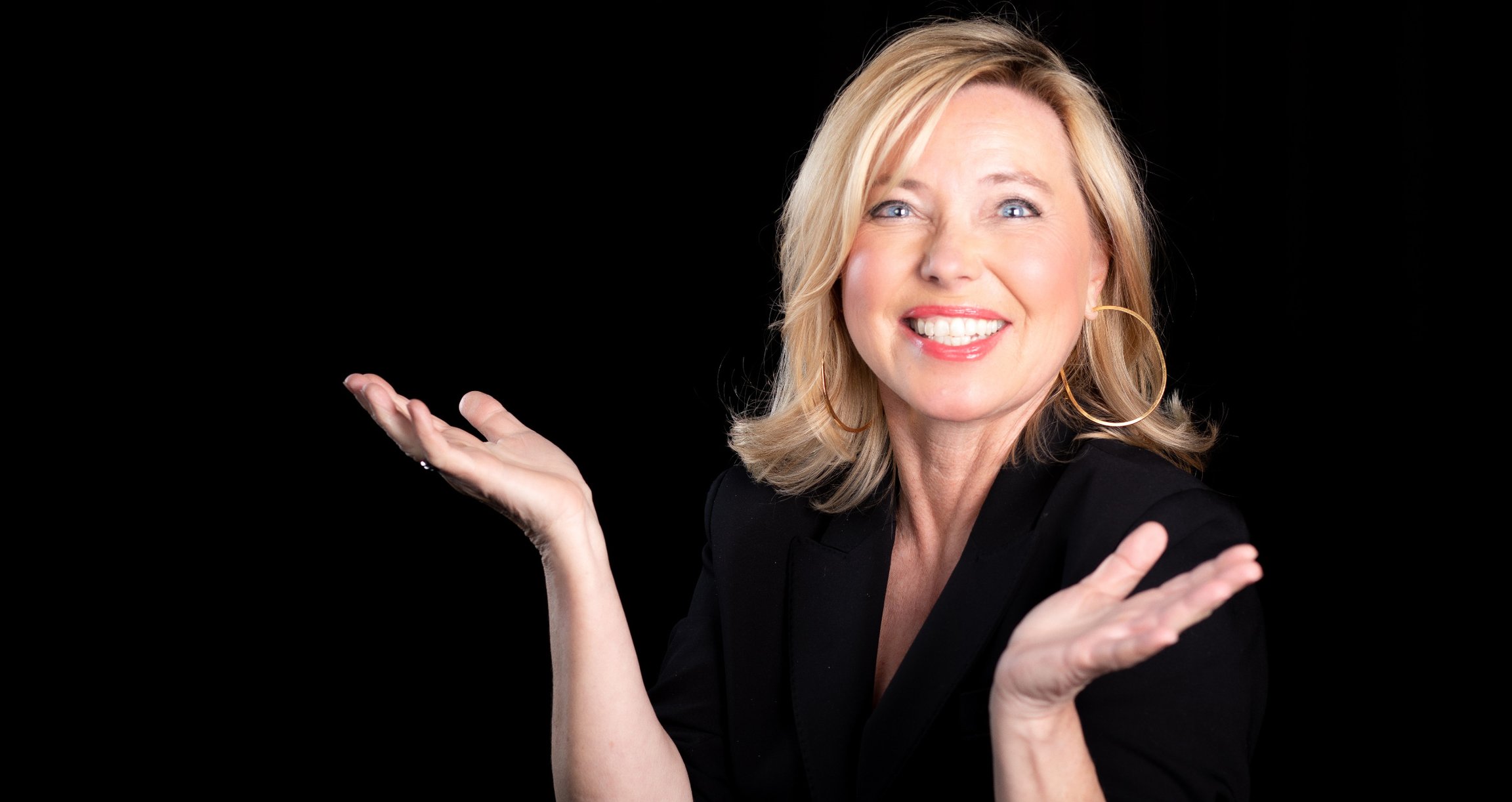 Blonde Dame in schwarzer Kleidung, lachend vor schwarzem Hinterrund