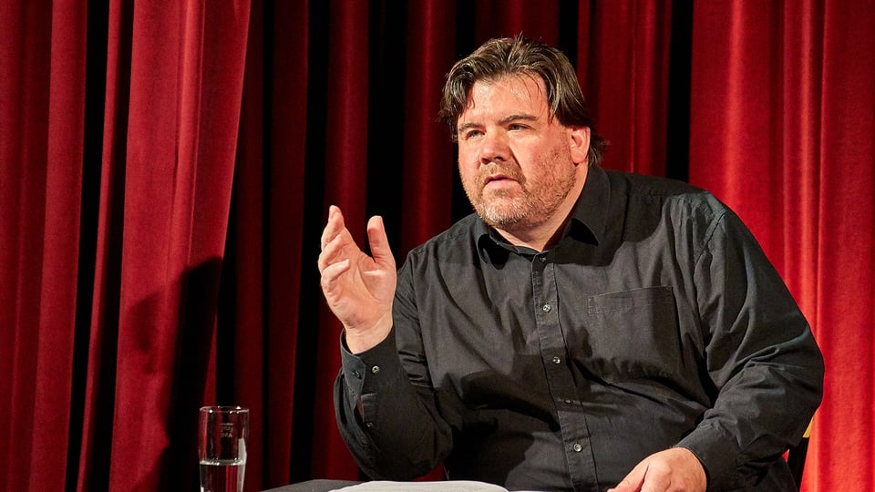 Ein Mann in schwarzem Hemd an einem Tisch sitzend, vor einem roten Vorhang