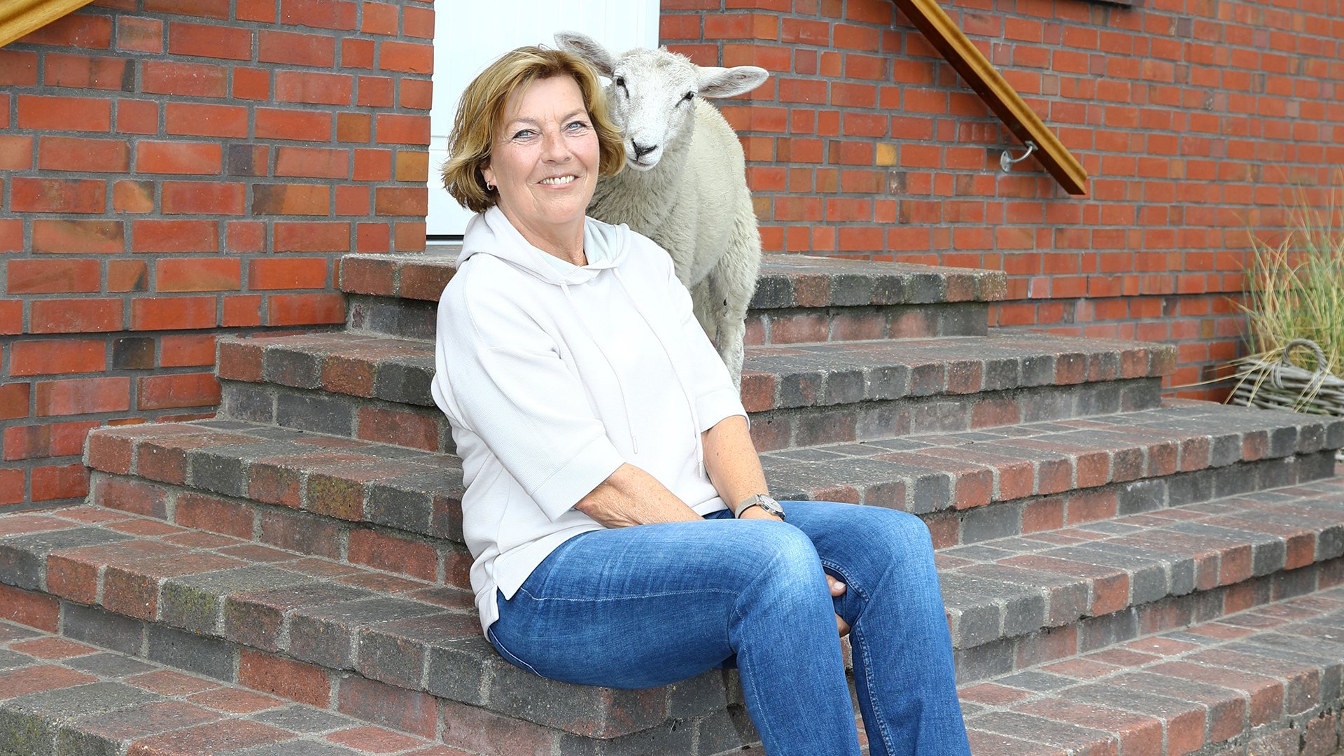 Die Autorin Dora Heldt lächelnd in weißer Bluse und Jeans sitzt an einer Ecke einer Treppe vor einer roten Ziegelwand, hinter ihr steht ein Schaf