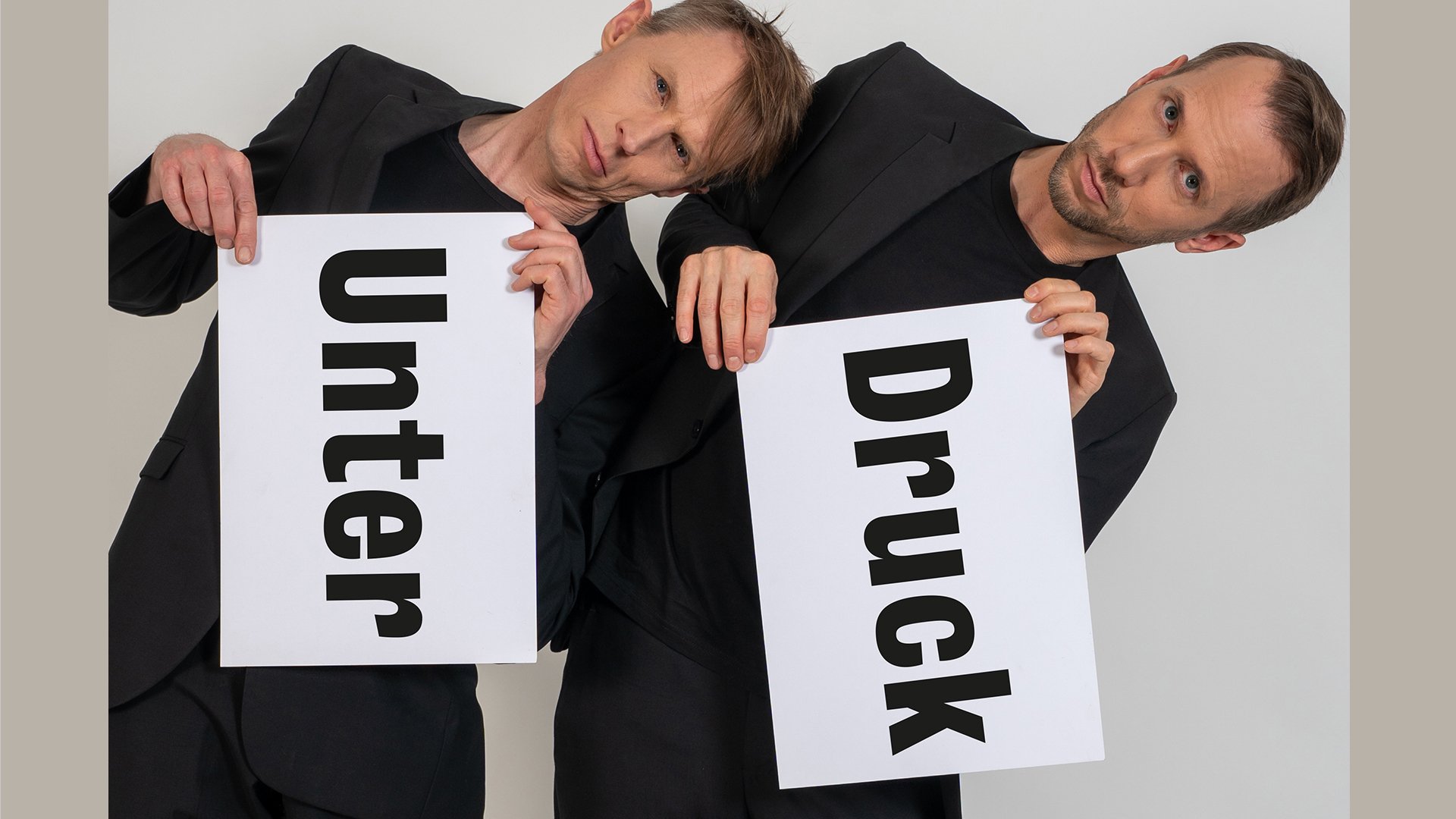 Foto von zwei Personen in schwarzen Outfits vor hellem Hintergrund, die weiße Tafeln mit den Aufschriften „Unter“ und „Druck“ halten und ihre Köpfe gegeneinander lehnen.