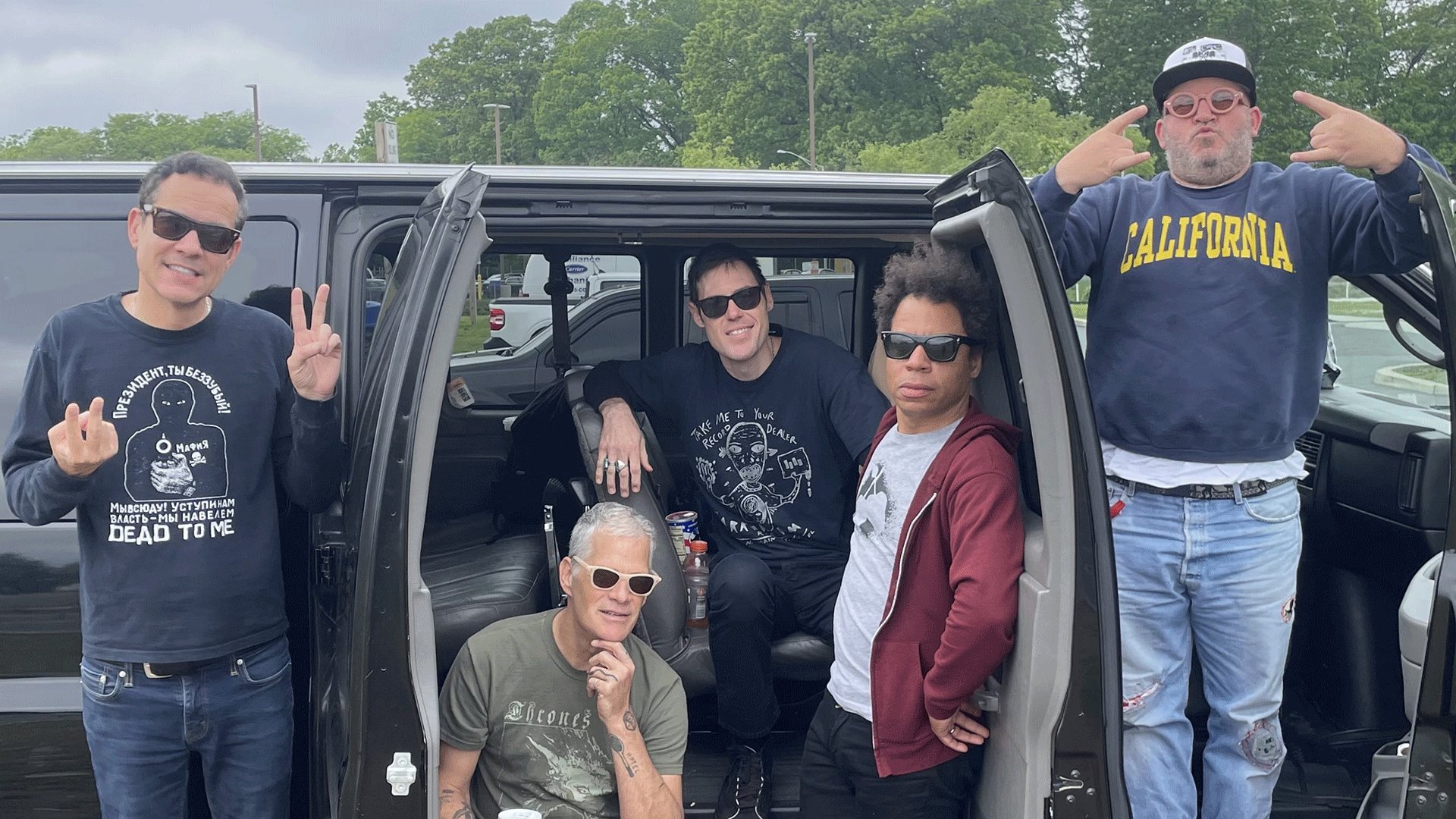 Bandfoto von Samiam mit fünf Bandmitgliedern vor einem geöffneten Van auf einem Parkplatz, vier Personen tragen Sonnenbrillen, eine Person steht mit erhobenen Händen neben dem Fahrzeug, im Hintergrund Bäume und bewölkter Himmel.