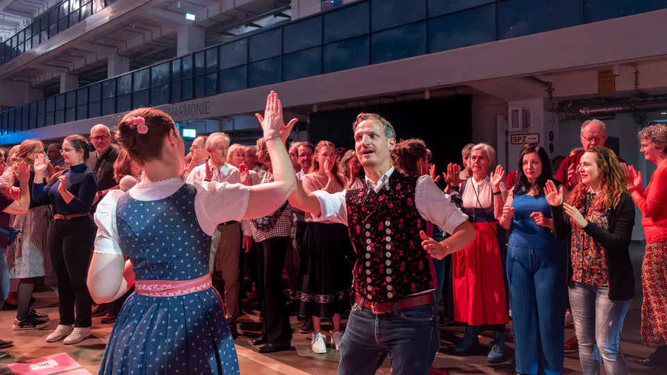 Gruppe von Menschen tanzt gemeinsam in einer Halle, Paar im Vordergrund in Tracht