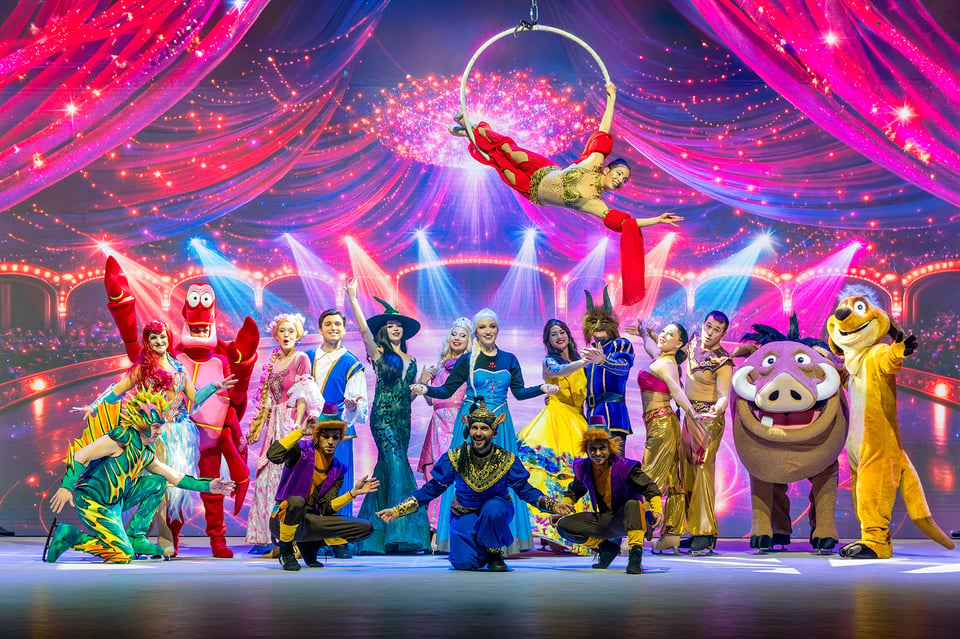 Bühnenfoto von Musical Dreams mit zahlreichen Darstellenden in farbenfrohen Kostümen bekannter Märchen- und Musicalfiguren, darunter eine Luftakrobatin am Ringtrapez, vor pink violett beleuchteter Showkulisse mit Lichteffekten