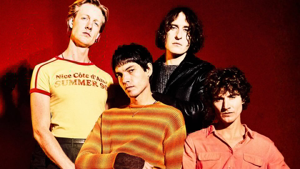 Vier Mitglieder der Band Temples stehen vor einem roten Hintergrund und blicken ernst in die Kamera; sie tragen farbige Retro-Outfits in Gelb, Orange und Rot sowie eine schwarze Lederjacke.