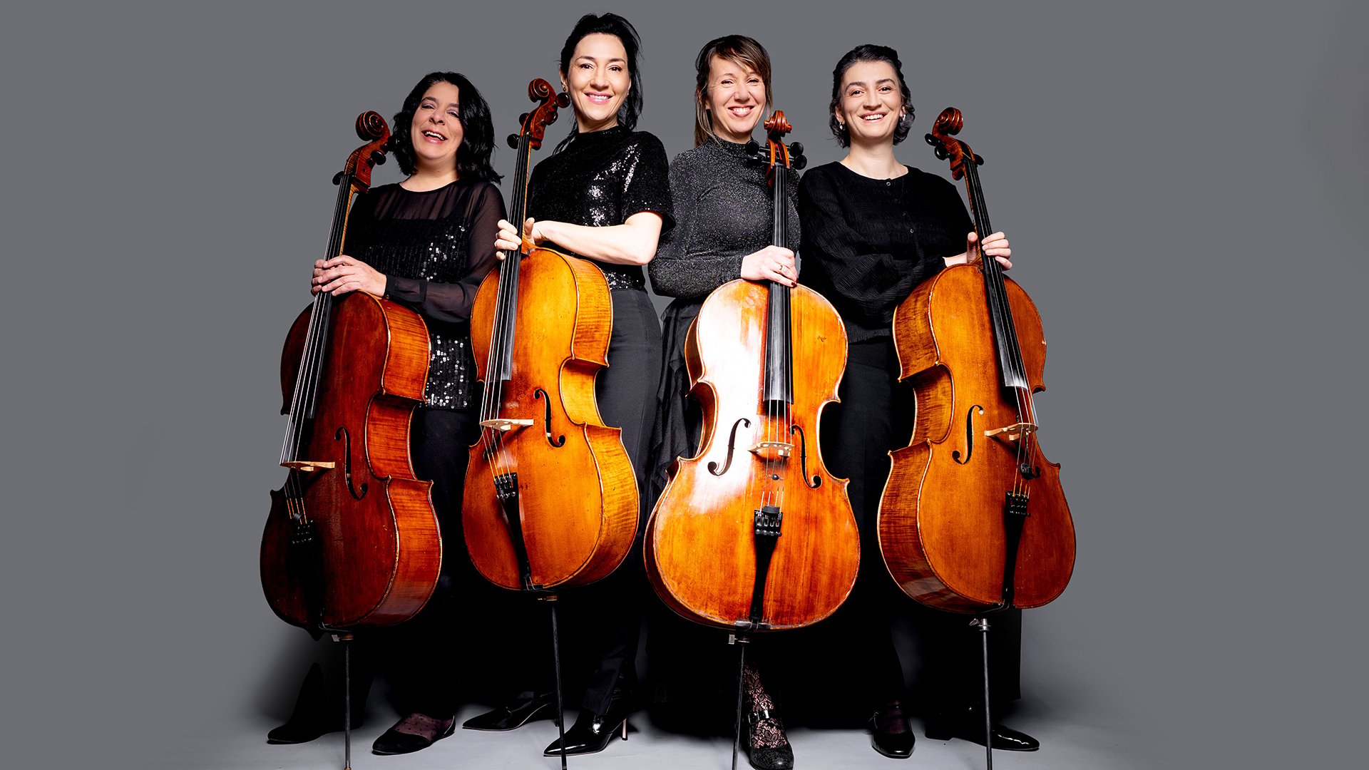 Vier Damen in dunkler Kleidun mit Cellos posieren lächelnd vor grauem Hintergrund