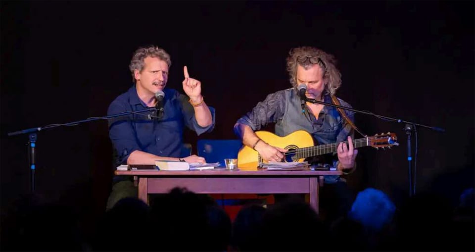 Foto von zwei Männern vor einem Tisch, beide mit Mikrofonen, einer von ihnen spielt Gitarre.