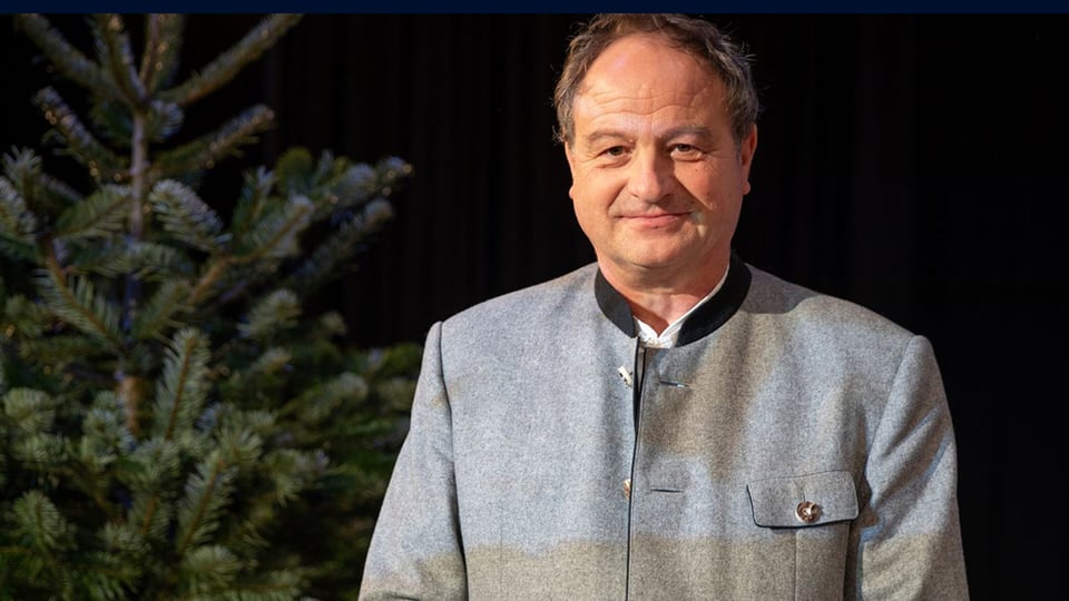 Rainer Maria Schiessler in grauer Trachtenjacke neben einem Tannenbaum vor dunklem Hintergrund