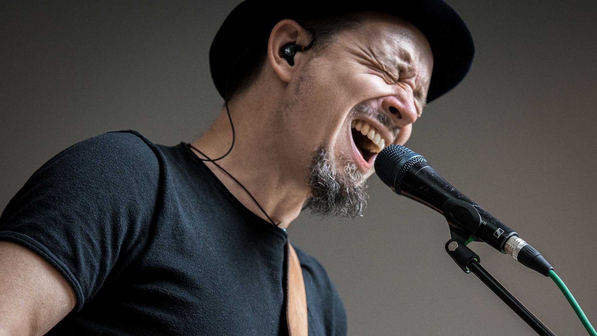 Der Künstler Tileema mit schwarzem Shirt, schwarzem Hut und Gitarre am Mikrofon vor dunkelgrauem Hintergrund