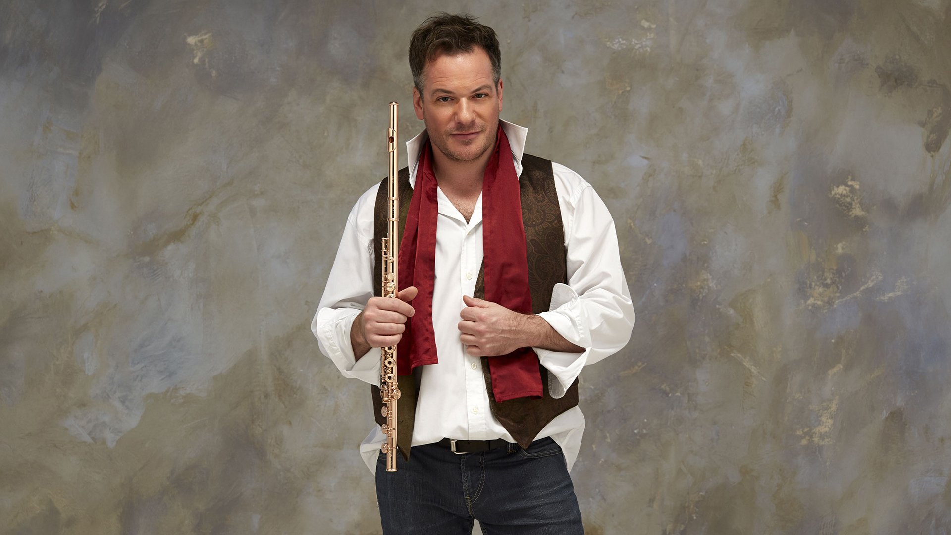 Emmanuel Pahud, der Soloflötist der Berliner Philharmoniker mit Querflöte in der Hand, weißem Hemd mit offenem Kragen, dunkle Weste roter Schal, marmorierter Hintergrund in Beige und Grau