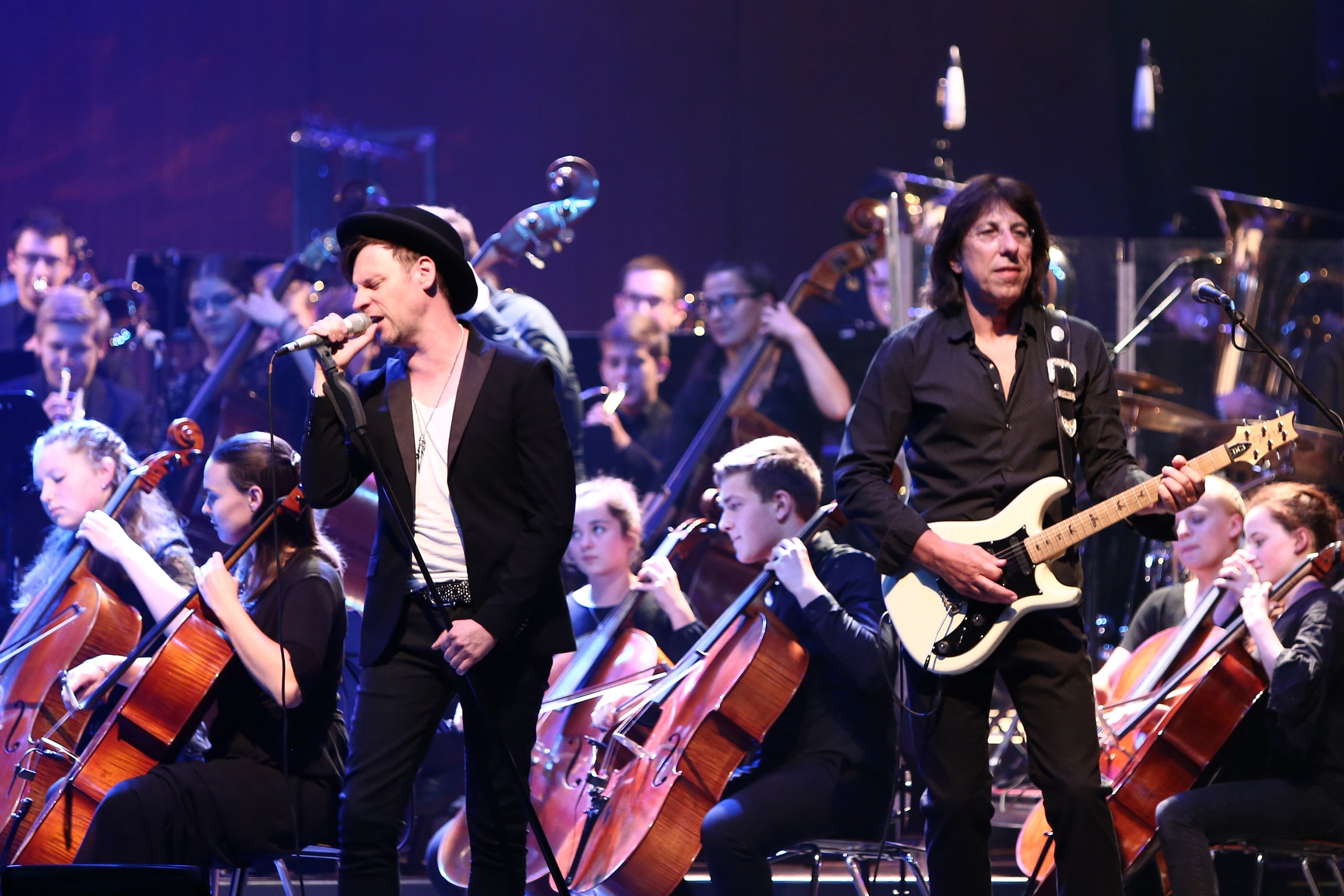 Zwei Musiker stehen auf einer Konzertbühne vor einem großen Orchester. Eine Person singt in ein Mikrofon, während die andere eine weiße E-Gitarre spielt. Im Hintergrund sind zahlreiche Streicherinnen und Streicher mit Celli und Kontrabässen zu sehen. Die Bühne ist in blau-violettes Licht getaucht und verbindet Rock- und Orchesterklänge in einer Live-Performance.