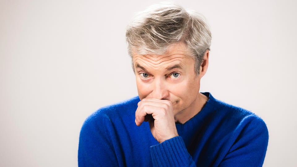 Portrait von Alfred Dorfer, der eine Hand vor den Mund hält, in blauem Pullover vor hellem Hintergrund
